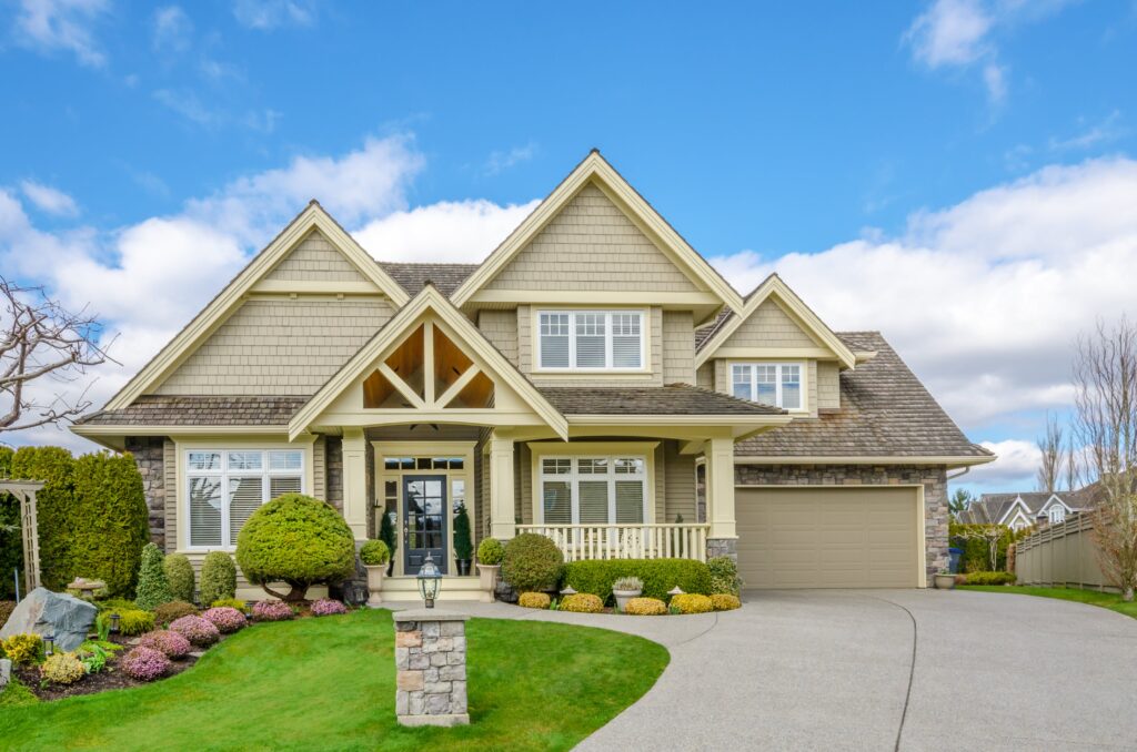 Two-story suburban house with beige siding, green landscaping, a covered front porch, and attached garage under a blue sky with clouds.