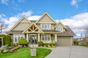 Two-story suburban house with beige siding, green landscaping, a covered front porch, and attached garage under a blue sky with clouds.