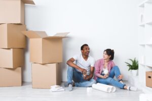 A man and woman sit on the floor among cardboard boxes, holding drinks, appearing to take a break while moving into a new home.
