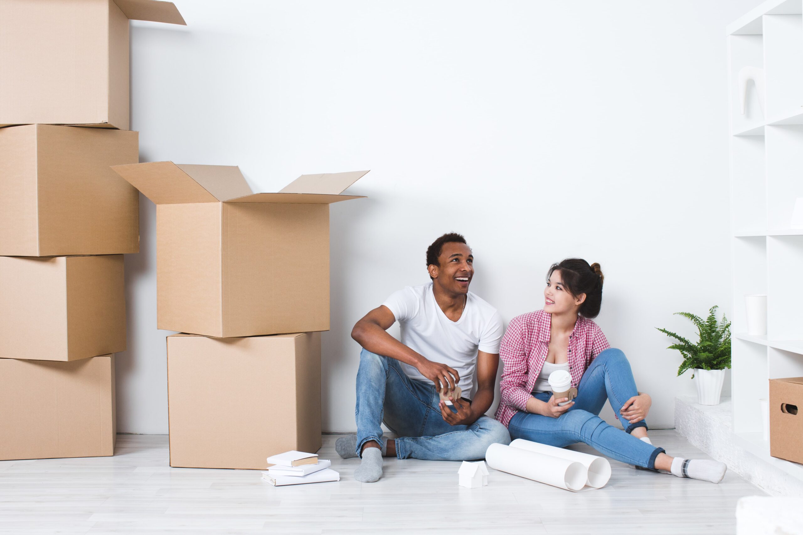 A man and woman sit on the floor among cardboard boxes, holding drinks, appearing to take a break while moving into a new home.