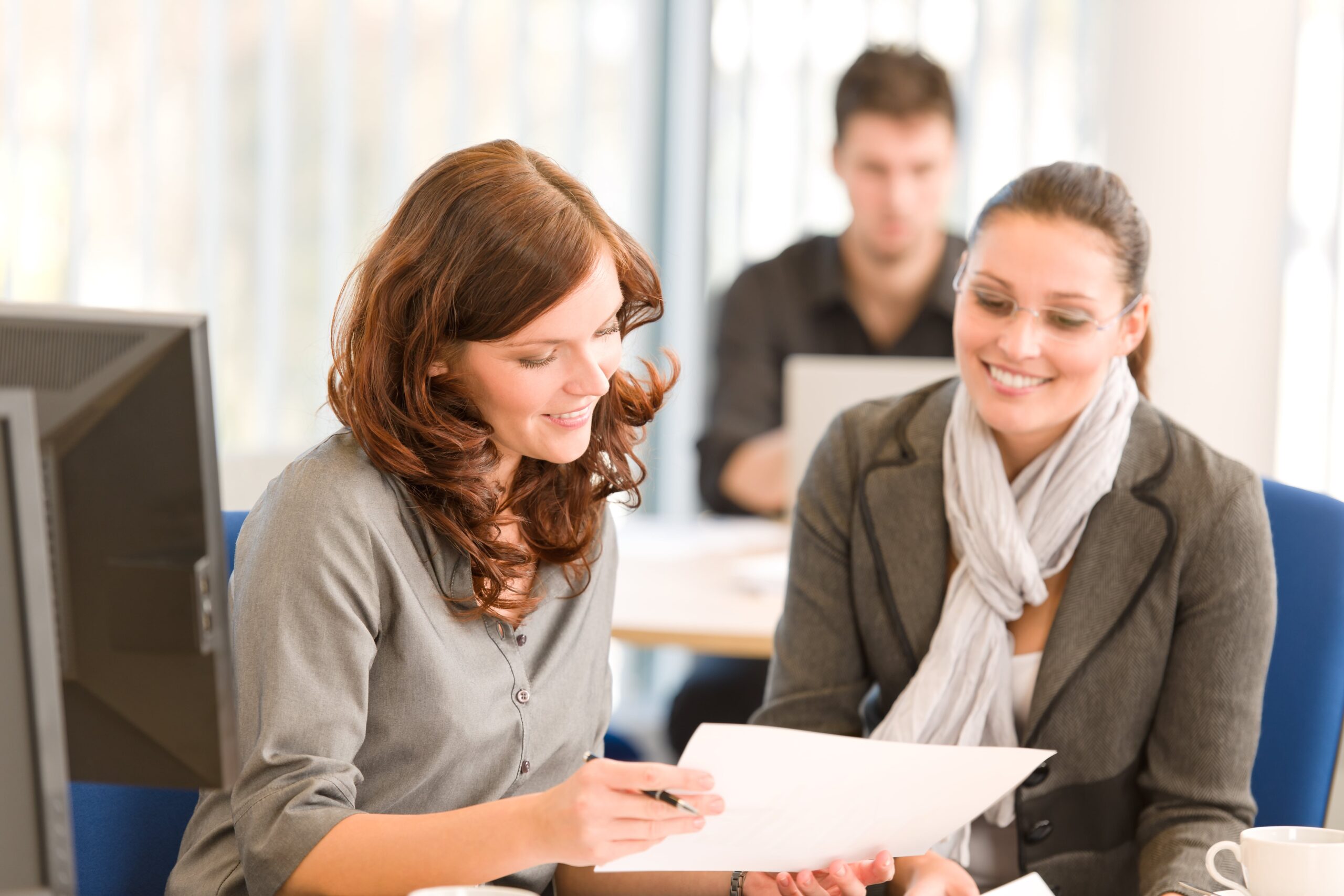 Two businesswomen seated at a desk, reviewing documents and discussing their work.