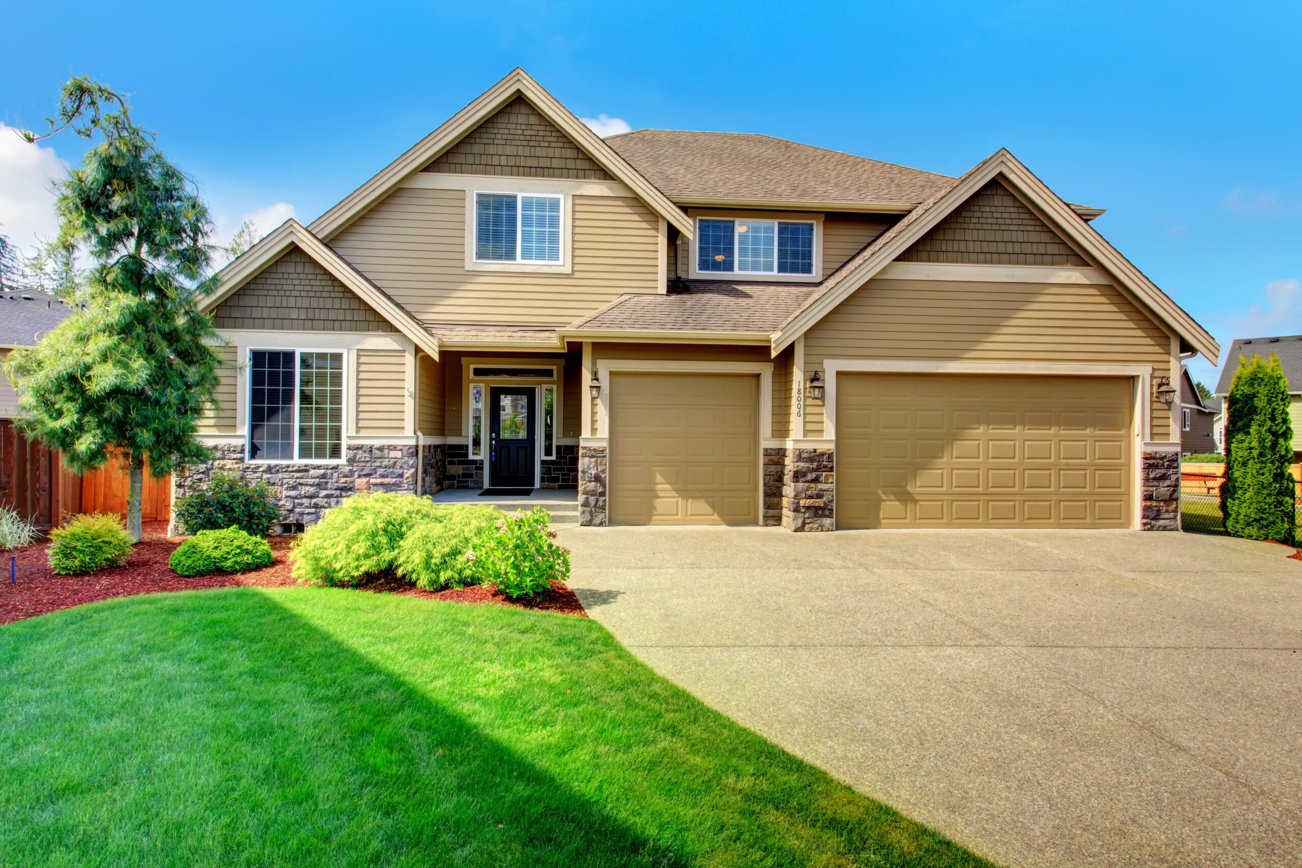 A spacious two-story house with beige siding and stone accents, featuring a large driveway and manicured lawn. A welcoming, suburban atmosphere.