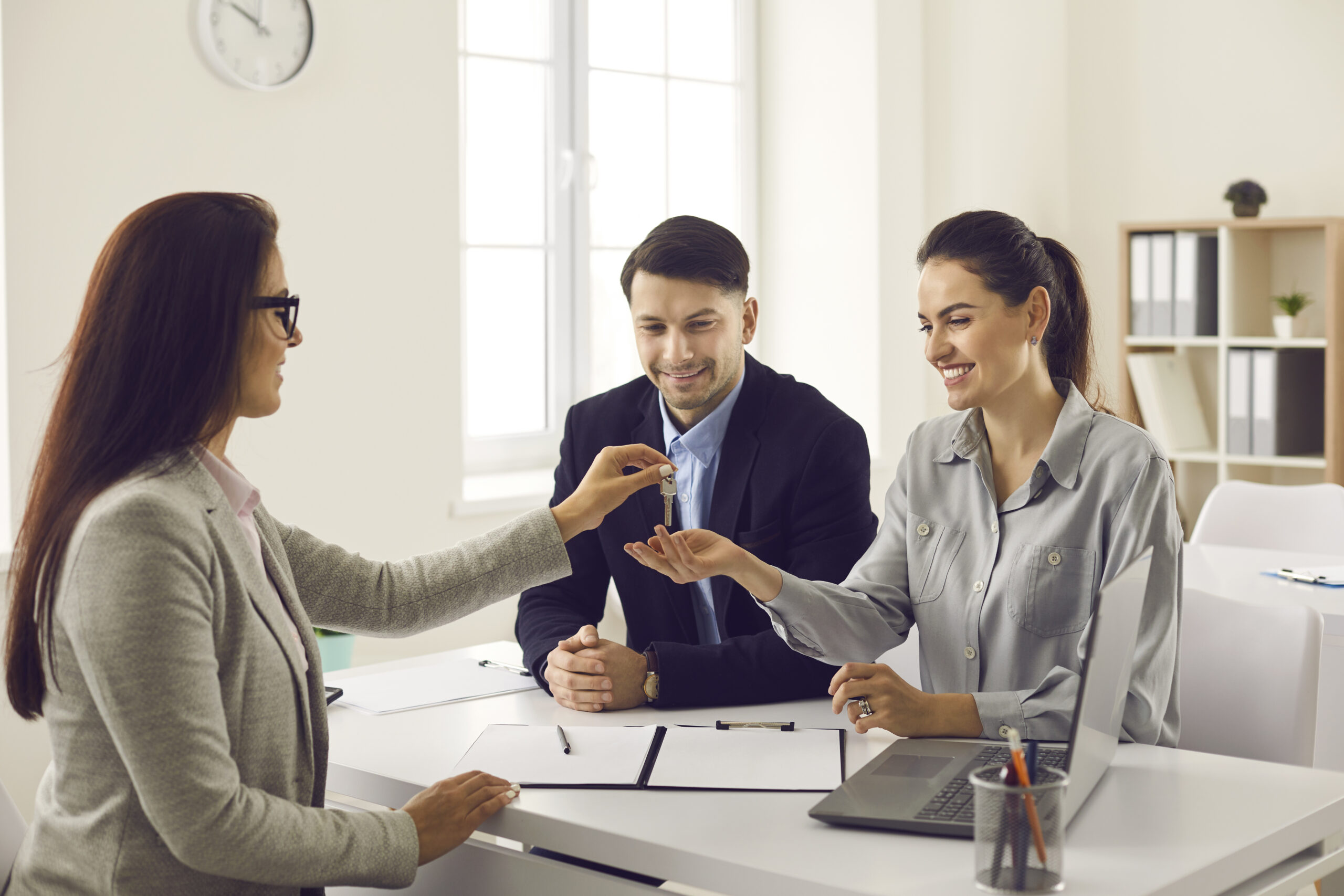 A woman in professional attire hands keys to a smiling couple across a desk. Papers and a laptop are on the table, suggesting a positive business exchange.