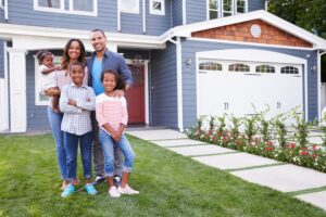 A smiling family of five stands together on a lush green lawn in front of a modern, blue two-story house with a white garage and blooming flowers.
