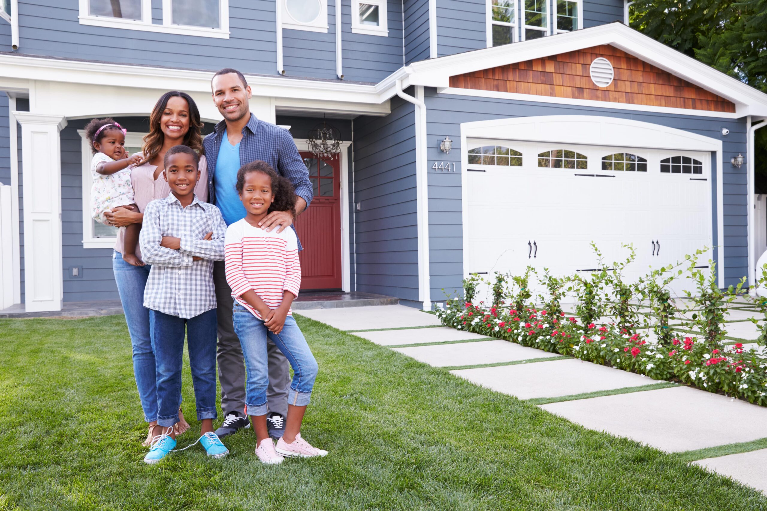 A smiling family of five stands together on a lush green lawn in front of a modern, blue two-story house with a white garage and blooming flowers.