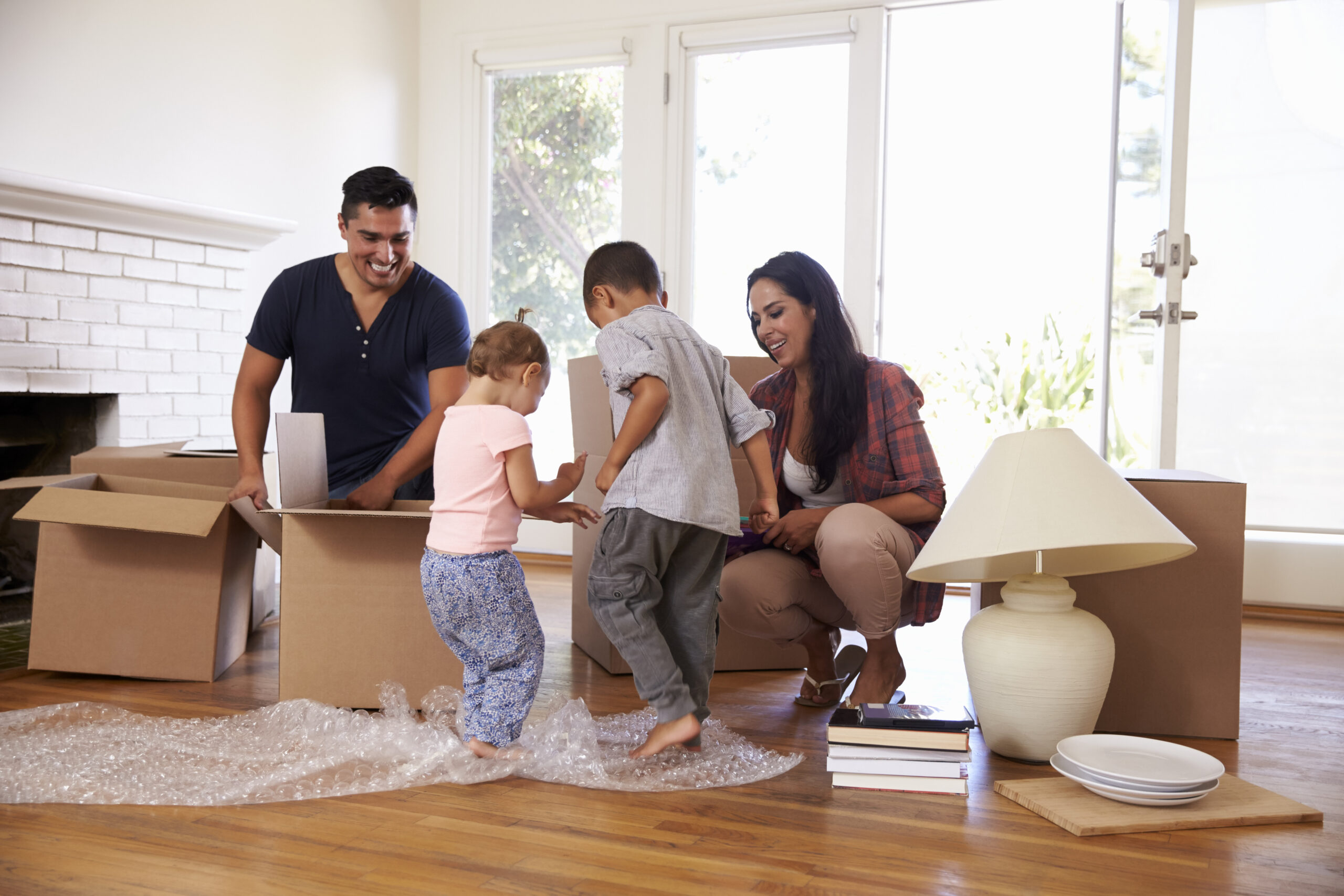 A family of four joyfully unpacks boxes in a sunlit room. The father smiles as children play on bubble wrap, while the mother crouches nearby, creating a warm, lively scene.