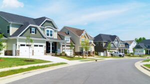 A row of modern suburban homes with well-maintained lawns and trees line a quiet, curved street under a clear blue sky, conveying a serene neighborhood vibe.