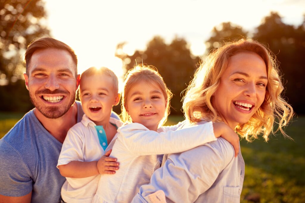 A joyful family of four smiles broadly in a sunlit park. The parents stand close, each holding a child. The scene conveys warmth and happiness.