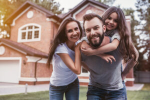 A cheerful family of three poses outside a brick house. The father carries the daughter on his back, while the mother stands beside them, all smiling warmly.